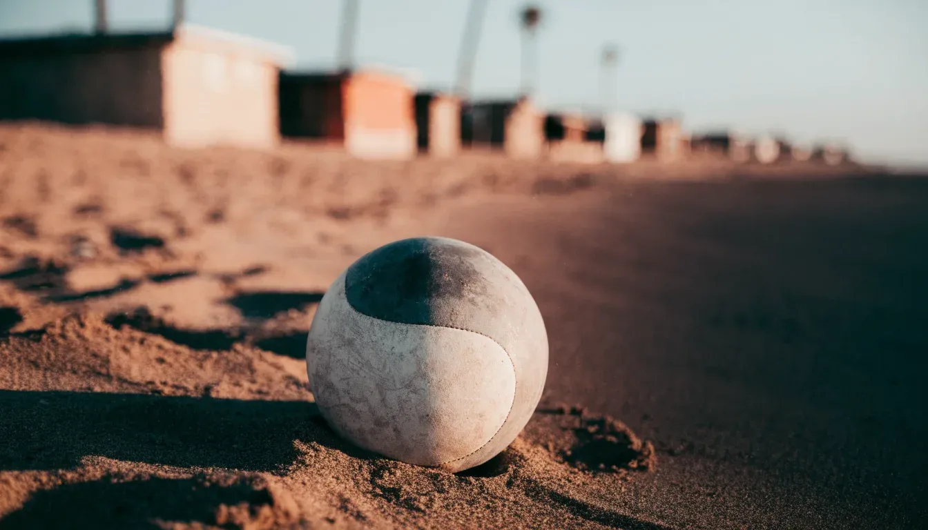 Ein Fussball liegt auf rotbraunem Sand am Rand eines Dorfes, die Abendsonne wirft lange Schatten.