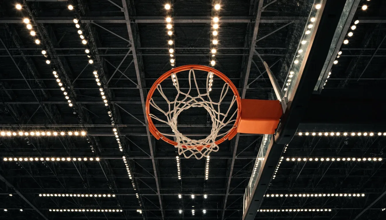 Ein Basketballkorb von unten gesehen, darüber eine Hallendecke mit Scheinwerfern und Stahlträgern.