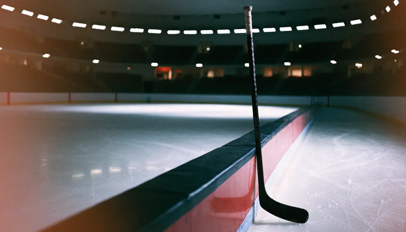 Ein Eishockeyschläger lehnt an der Bande, in der Ferne liegt die hell beleuchtete Eisfläche einer Halle unter Flutlicht.