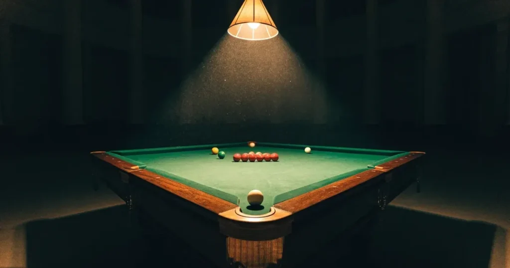 Ein Snookertisch mit grünem Tuch und einem Dreieck aus roten Kugeln unter einer tief hängenden Lampe in einer leeren Halle.