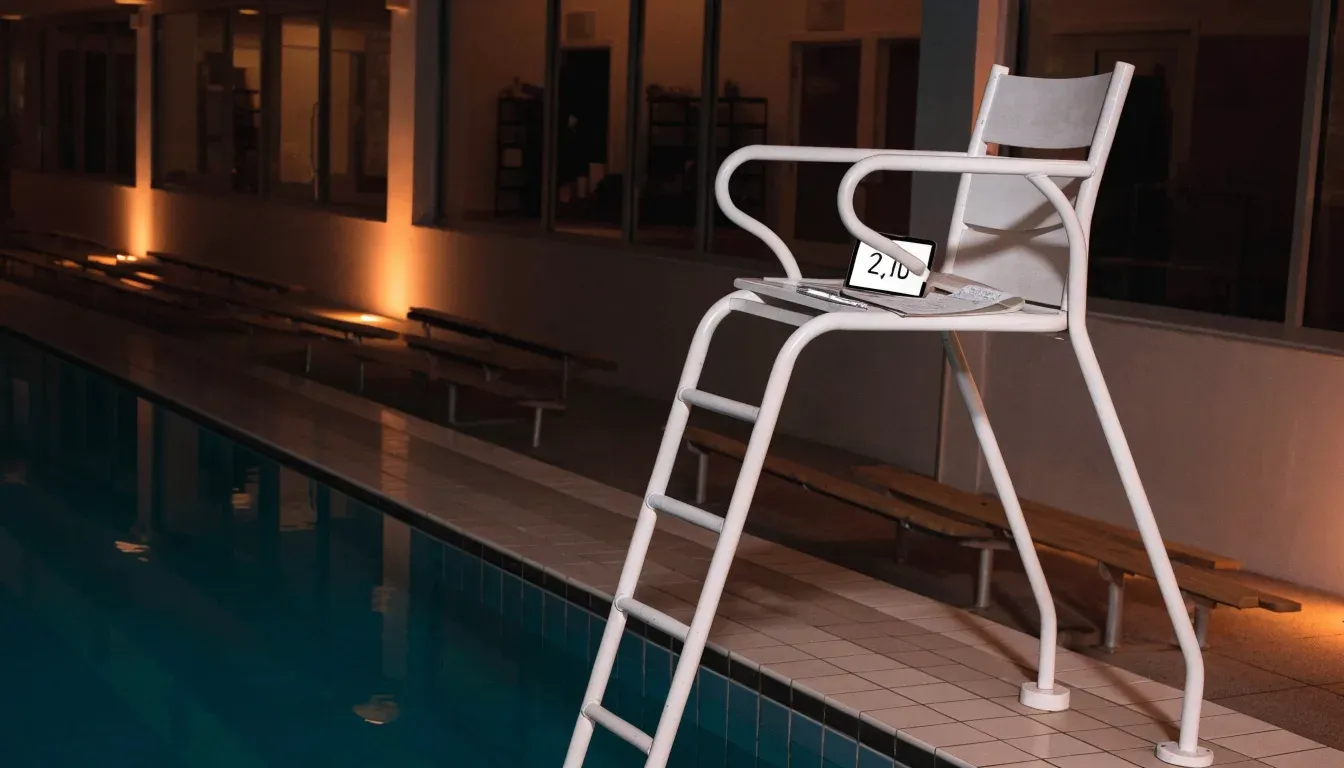 Ein Bademeisterstuhl neben einem Hallenbad zur Schliesszeit, gedimmtes Licht, glatte Wasseroberfläche.