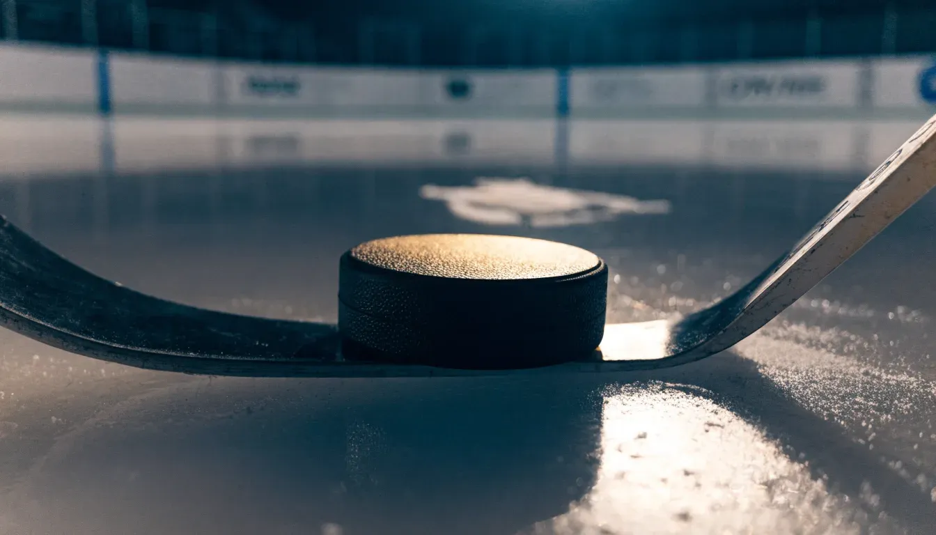 Ein Eishockeypuck und ein Schläger liegen auf einer glänzenden Eisfläche, im Hintergrund verschwimmen leere Banden.